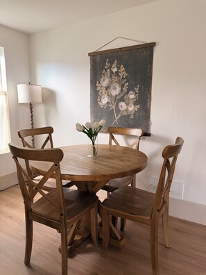 A round wooden dining table with four chairs, a vase with flowers, and a wall-mounted painting.