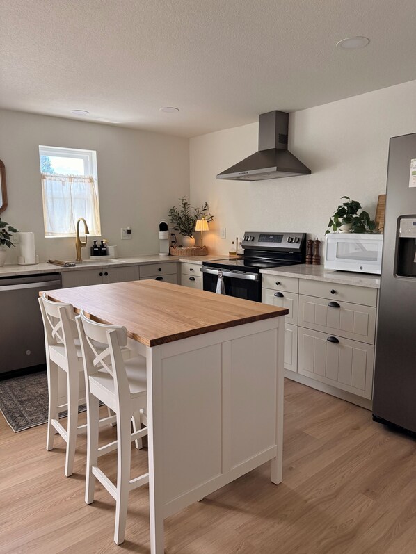 A modern kitchen with a wooden island, white cabinetry, stainless steel appliances, and a window with sheer curtains.