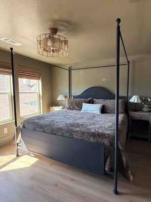 A four-poster bed with a dark headboard, a bedside table with a lamp, and a window with blinds.