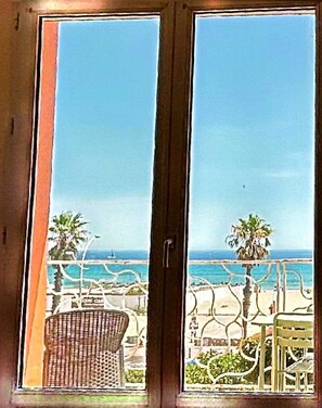 Vista a través de una ventana con marco de madera, que da a una playa con palmeras y un cielo despejado.
