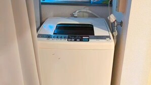 Una lavatrice compatta e bianca, portatile, con pannello di controllo digitale, posizionata in un angolo con una finestra sopra.