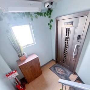 A room with a wooden desk, a fire extinguisher, a window, a door with a number plate, and a security camera. - Youki Residence (Suginami-ku)