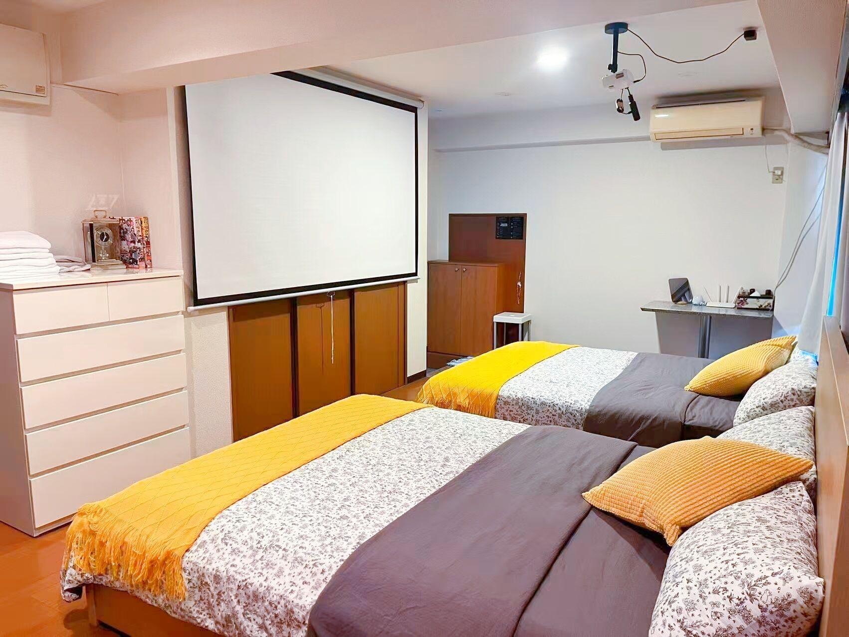 A hotel room with two beds, a desk, a wardrobe, and a projector screen.