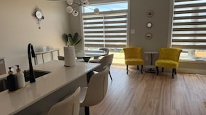 A modern kitchen with a central island, a dining area with yellow chairs, and large windows with blinds.