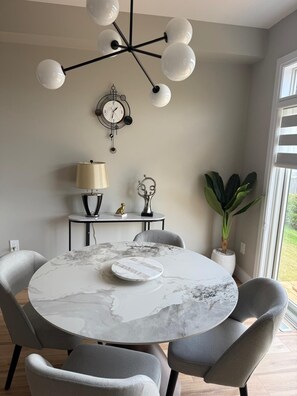 A dining area with a round marble table, grey upholstered chairs, a wall-mounted clock, a table lamp, and a potted plant. - 5 Bedrooms deluxe, Hyde Park Mall and Western University area Sleep up to 10 (London)