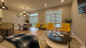 A modern living room with a sofa, a coffee table, and yellow chairs. There is a dining area with a table and chairs, and a large window with blinds. - 5 Bedrooms deluxe, Hyde Park Mall and Western University area Sleep up to 10 (London)