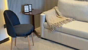 Una habitación con una silla de color oscuro, un sofá de tonos claros, una mesita redonda con dos cuadros enmarcados y una pared con textura.