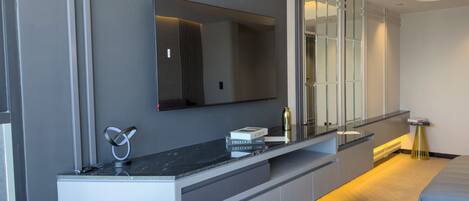 A modern hotel room with a large flat-screen TV mounted on the wall, a built-in console table with books and a water bottle, a sofa, and a bedside table with a lamp.