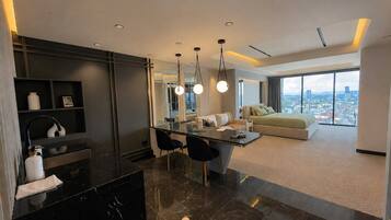 A modern hotel room with a dining area, a sofa, and a large window offering a city view.