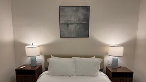 Una habitación de hotel con una cama, dos mesitas de noche con lámparas, una obra de arte en la pared y una paleta de colores neutros.