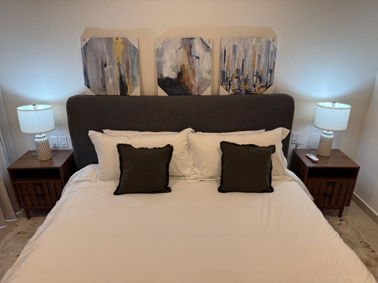 Un lit avec une literie blanche, deux oreillers foncés et une tête de lit sombre. Il y a deux tables de chevet avec des lampes et une œuvre d’art accrochée au mur au-dessus du lit.