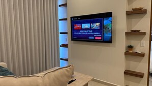 Una sala de estar con una televisión de pantalla plana montada en la pared, un sofá, una mesa de centro y un ventanal con persianas verticales.