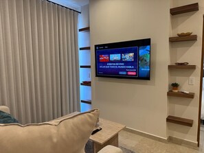 A living room with a flat-screen TV mounted on the wall, a sofa, a coffee table, and a window with vertical blinds. - Vista Cortés Ocean View | Walk to the Malecón (La Paz)
