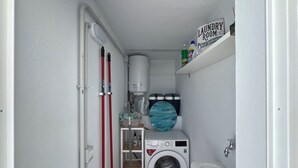 A compact laundry room with a washing machine, a tub, and a shelf with cleaning supplies. - Casita Coco – Your Lanzarote Home for Longer Stays | Work, Relax, Explore (Guatiza)