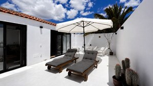 A modern outdoor patio with two lounge chairs, a large umbrella, and potted cacti. - Casita Coco – Your Lanzarote Home for Longer Stays | Work, Relax, Explore (Guatiza)