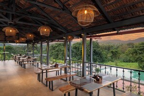 An outdoor dining area with tables and benches, surrounded by a railing, overlooking a lush landscape with a view of distant mountains.