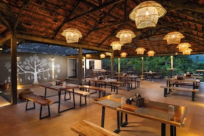 An outdoor dining area with multiple tables and benches, surrounded by a thatched roof structure.