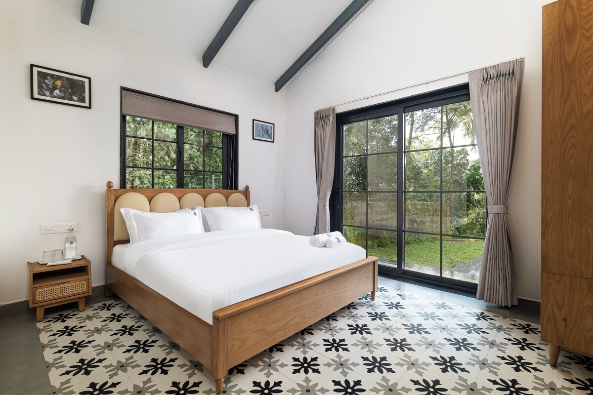 A bedroom with a wooden bed, a bedside table, a window with curtains, and a view of greenery outside.