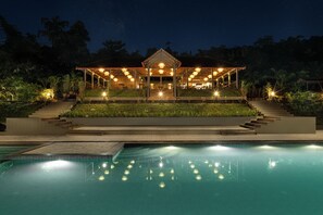 Vista nocturna de un resort con piscina, un área exterior techada con asientos y una entrada bien iluminada.