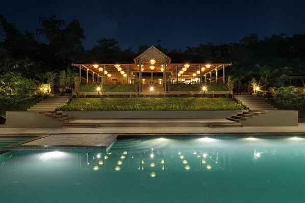A nighttime view of a resort with a pool, a covered outdoor area with seating, and a well-lit entrance.