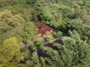 An aerial view of a property surrounded by dense greenery, featuring a central building with a red roof, a paved driveway, and a small garden area.