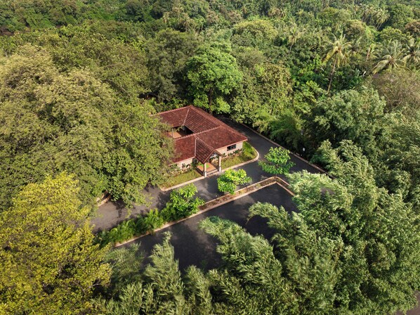 An aerial view of a property surrounded by dense greenery, featuring a central building with a red roof, a paved driveway, and a small garden area.