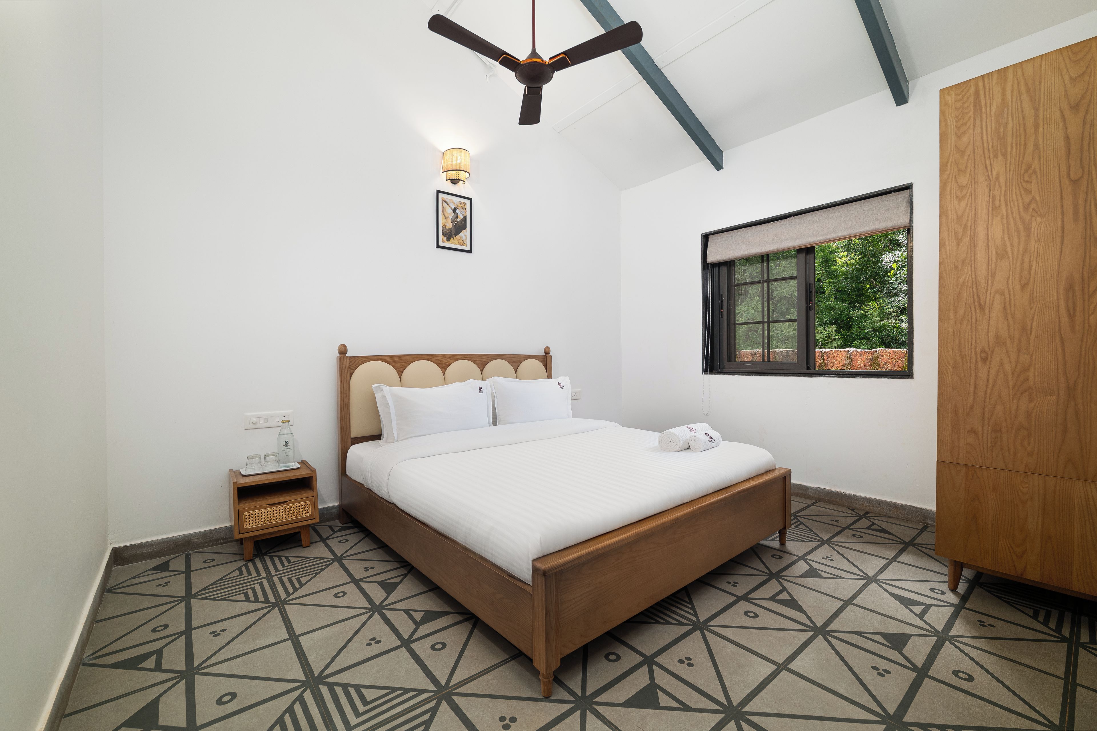 A bedroom with a bed, bedside table, nightstand, wall-mounted lamp, and a window with a view of greenery.