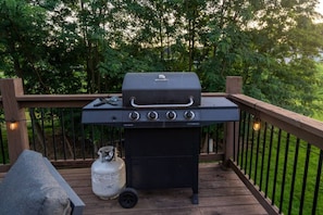 Ein Grill mit Holzkohle auf einer Holzterrasse, mit einer Gasflasche und einem Schubkarren in der Nähe.