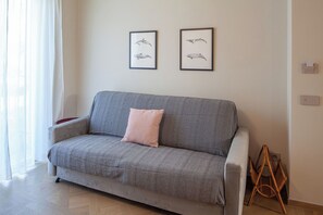 Ein graues Sofa mit einem rosafarbenen Kissen, zwei gerahmte Tierillustrationen an der Wand und ein Korbstuhl.