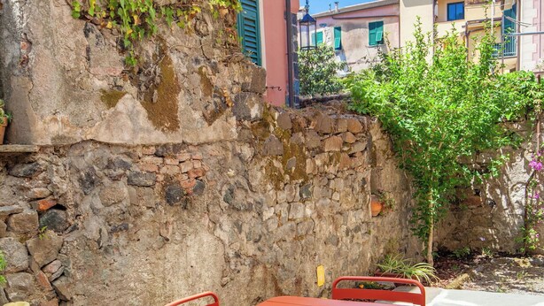 Uma área externa com mesas e cadeiras, uma parede de pedra e vista para edifícios ao fundo.