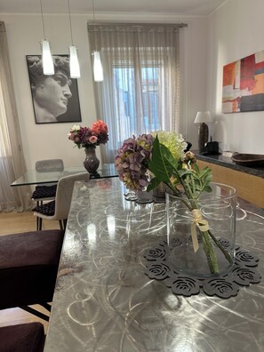 Uma sala de jantar com mesa de vidro, cadeiras, um vaso com flores, um quadro na parede e luminárias pendentes.