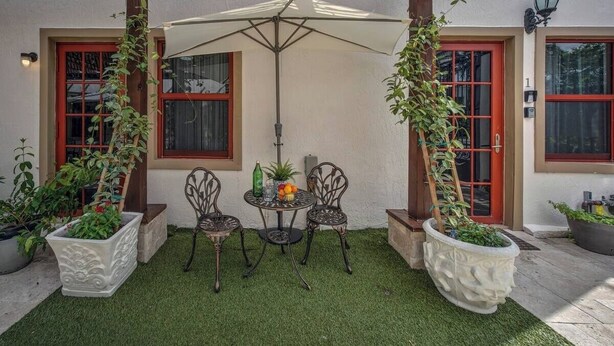Un patio con césped artificial, dos sillas de metal, una mesa con sombrilla blanca y plantas en macetas.
