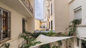 Un estrecho patio con un balcón, plantas en macetas y un dosel blanco.