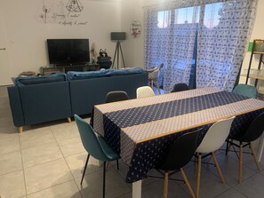 A living room with a sofa, a TV, a dining table with chairs, and a patterned tablecloth. - 3-bedroom house with a pool, in a quiet neighborhood (Saint-Jean-de-Monts)