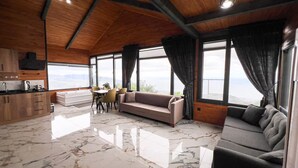 A spacious living area with a sofa, a dining table with chairs, a kitchen area, and large windows offering a view. - Trabzon’da Jakuzili Deniz, Dağ Manzaralı Bungalov (Akçaabat)