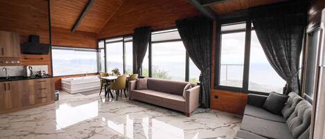 A spacious living area with a sofa, a dining table with chairs, a kitchen area, and large windows offering a view.