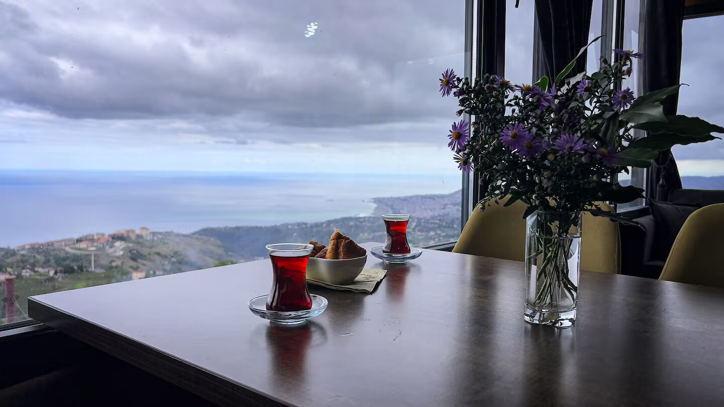 A table with a view of a coastal landscape, a vase with flowers, and two cups of tea.