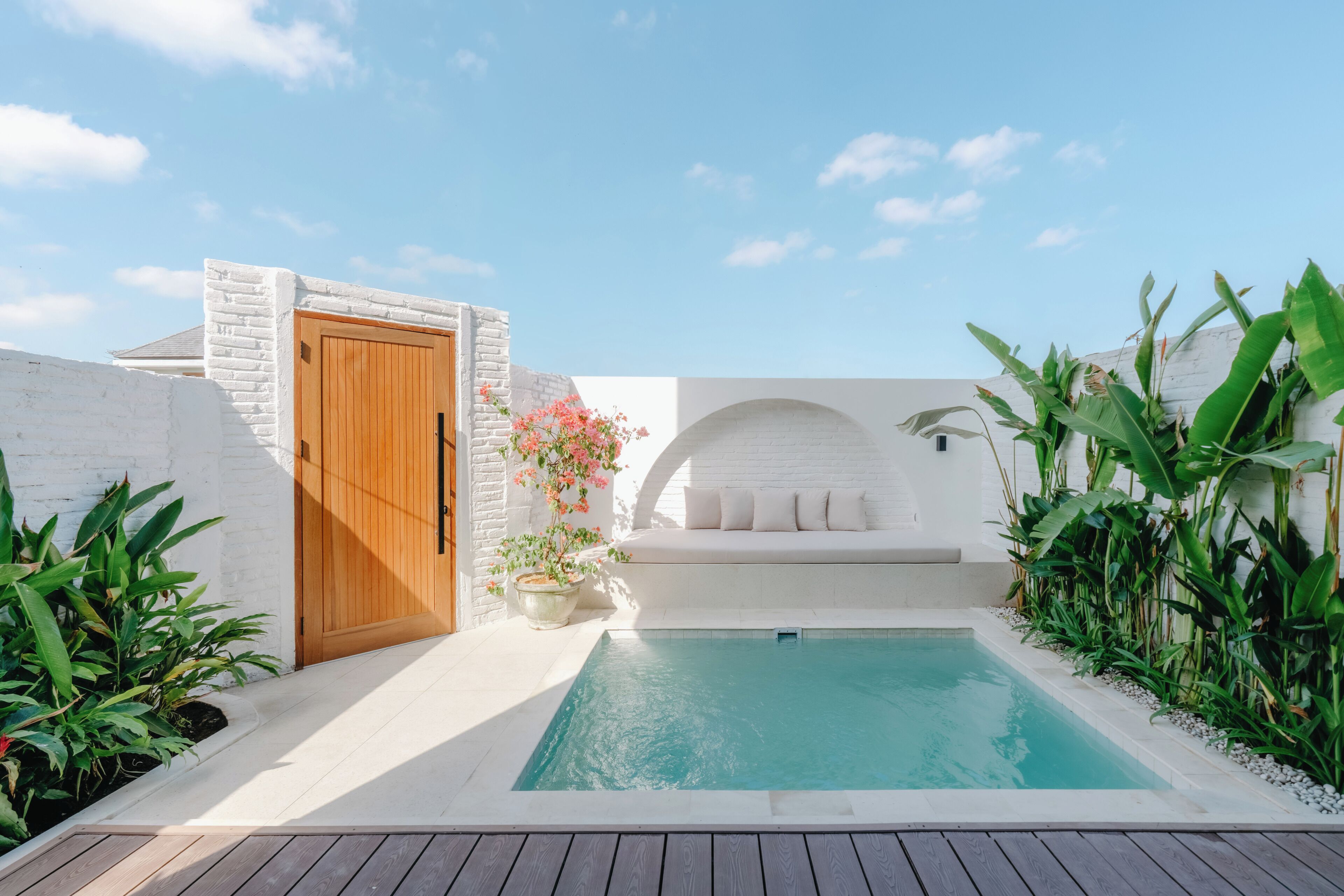 Une piscine avec une porte en bois, des murs en briques blanches, un banc blanc et des plantes vertes.
