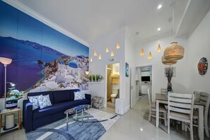 A modern living room with a large mural, a sofa, a coffee table, and a dining area with a chandelier. - Santorini Patio Suite 1Bed/2Bath/1K - DT - Hot Tub (Fort Lauderdale)