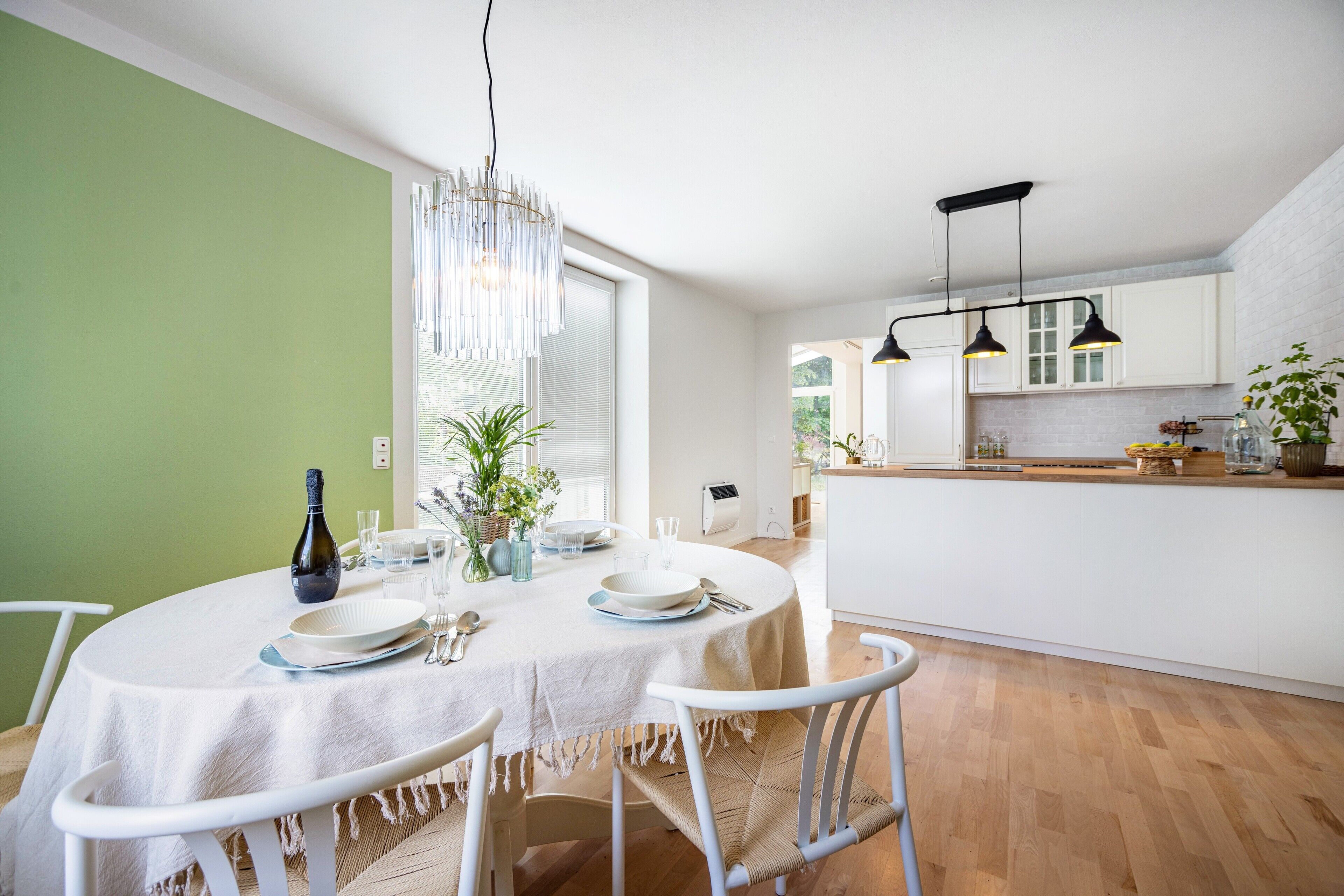 Een eetgedeelte met een ronde tafel, witte stoelen, een kroonluchter en een keuken op de achtergrond.