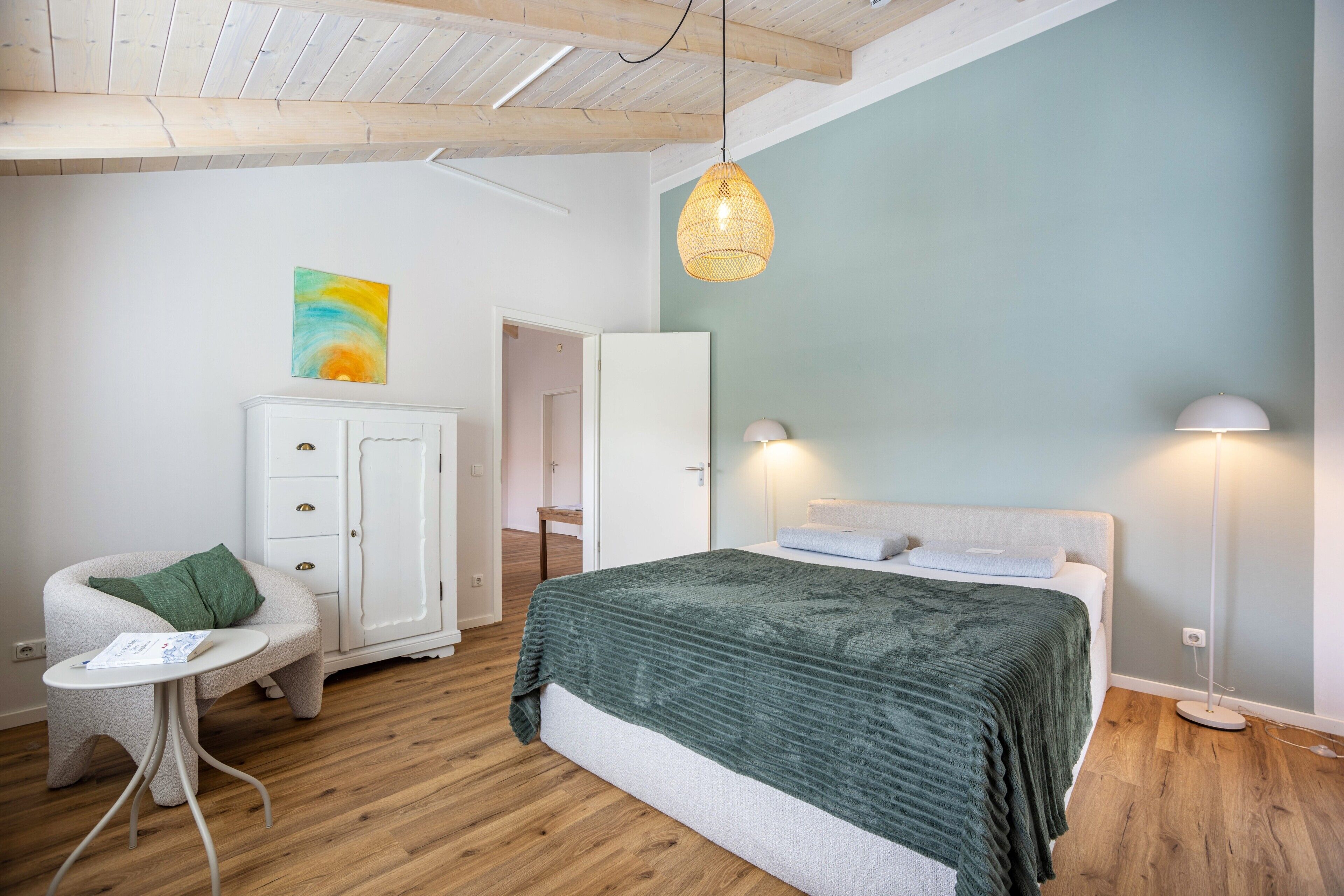 A bedroom with a bed, a chair, a small table, a wardrobe, and a painting on the wall.