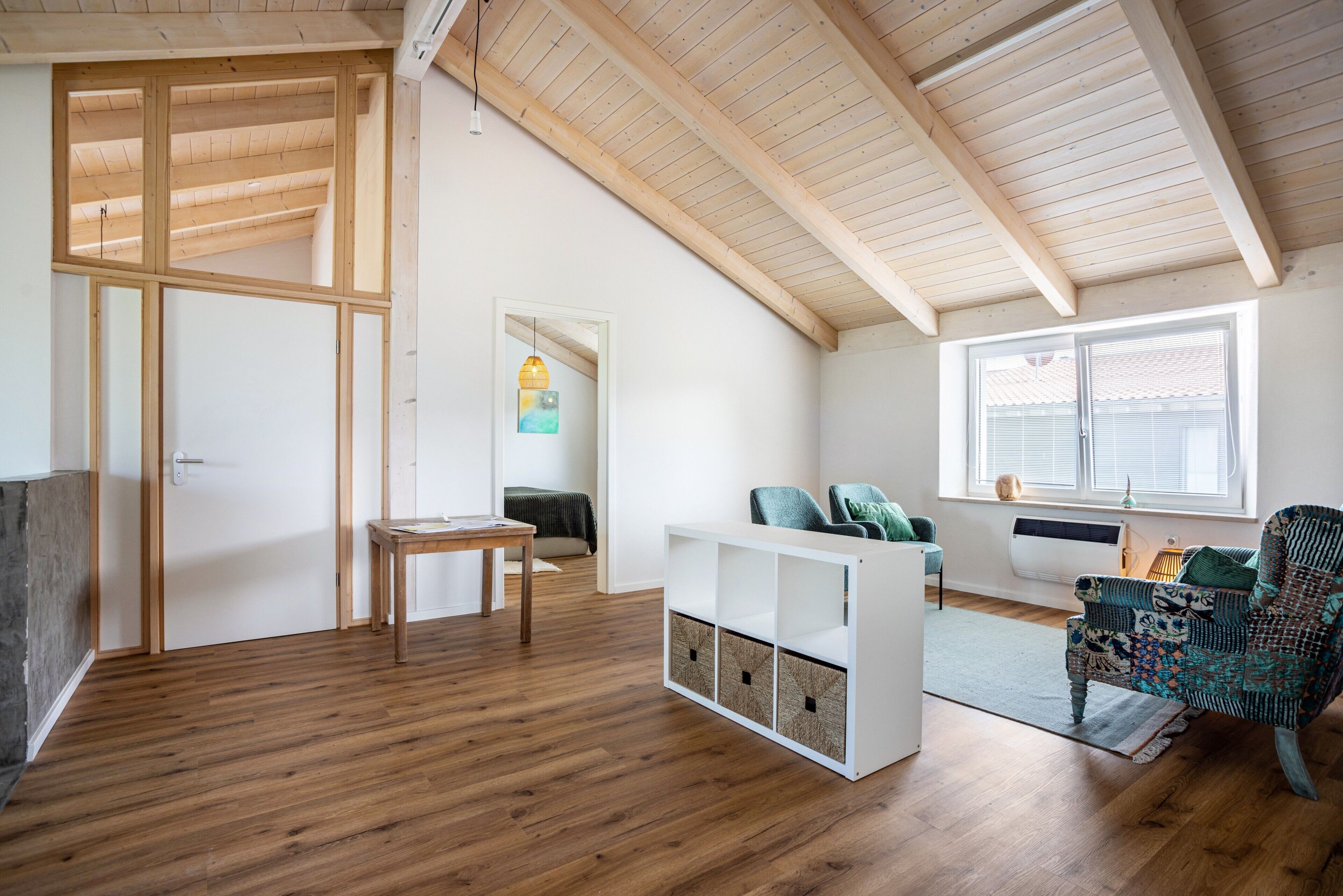 Ruime kamer met schuine plafonds, houten vloer, witte muur, zithoek en een gestreept fauteuil, plus een tafeltje.