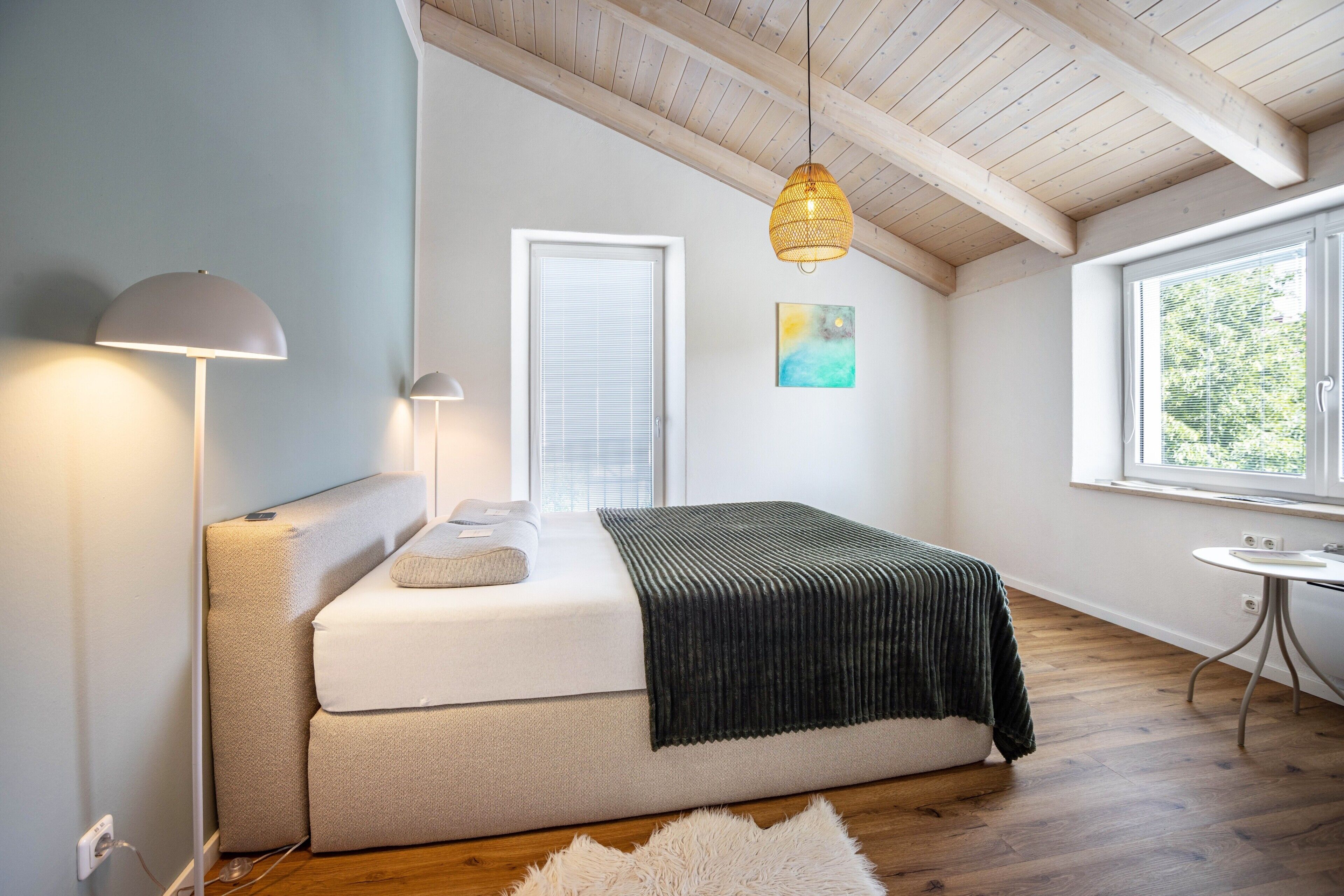 Ein Schlafzimmer mit einem Bett, zwei Nachttischlampen, einem Fenster mit Jalousien, einem Gemälde und einem kleinen Tisch.