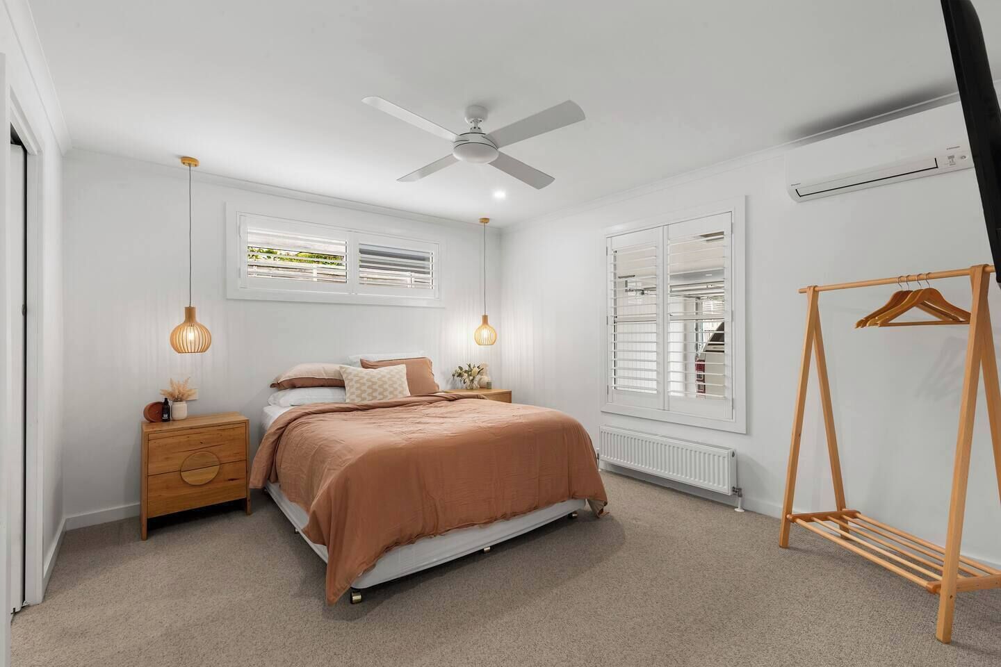 A bedroom with a bed, a ceiling fan, a radiator, a wooden dresser, a hanging light fixture, and a wooden clothes rack.