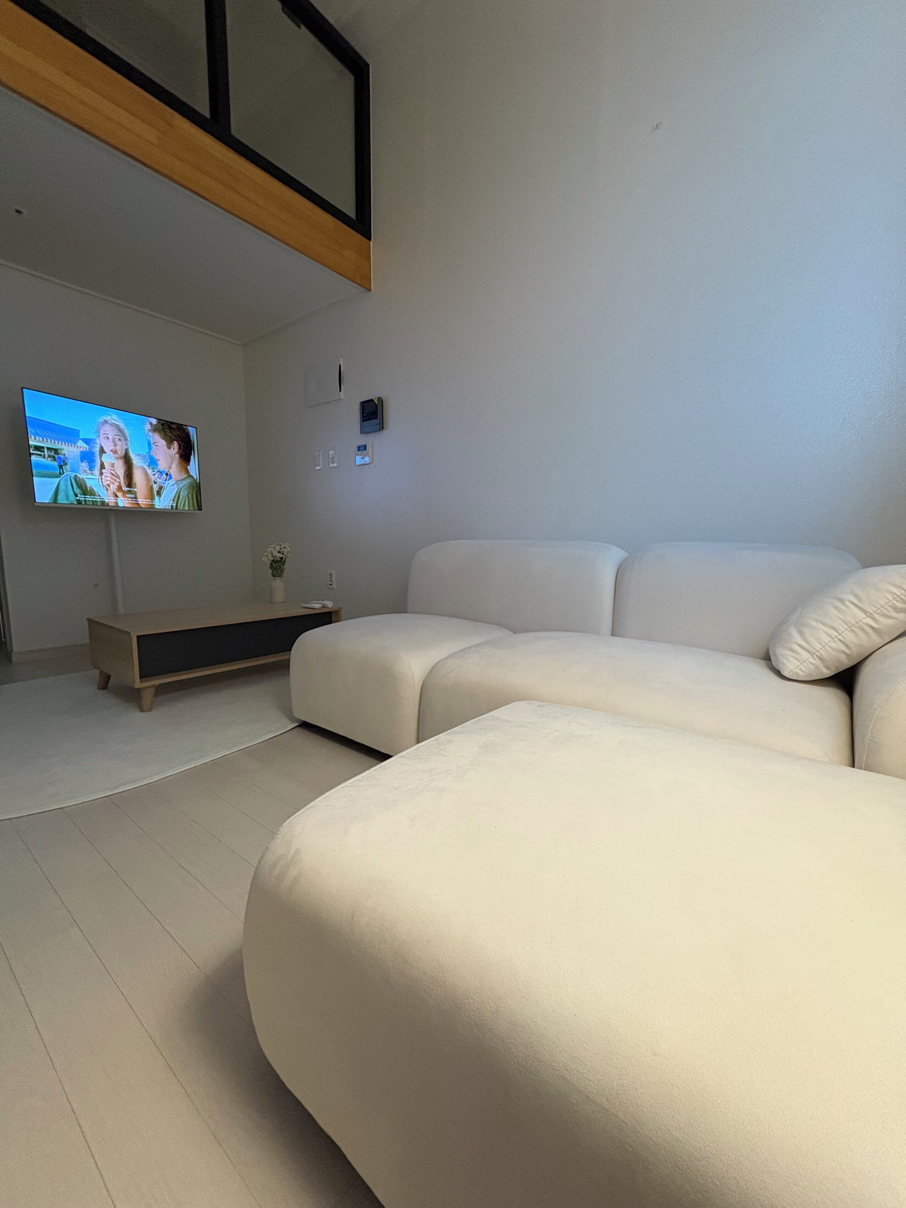 Un salon moderne avec un grand canapé d’angle blanc, un téléviseur à écran plat fixé au mur et une table basse en bois au design minimaliste.