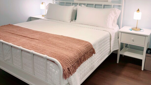 A neatly made bed with a white and brown blanket, a bedside table with a lamp, and a window with blinds.