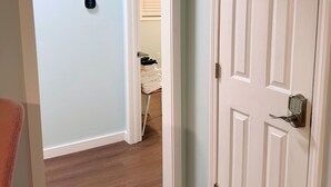 A hallway with a white door, a thermostat, and a glimpse of a room with a chair and a window.