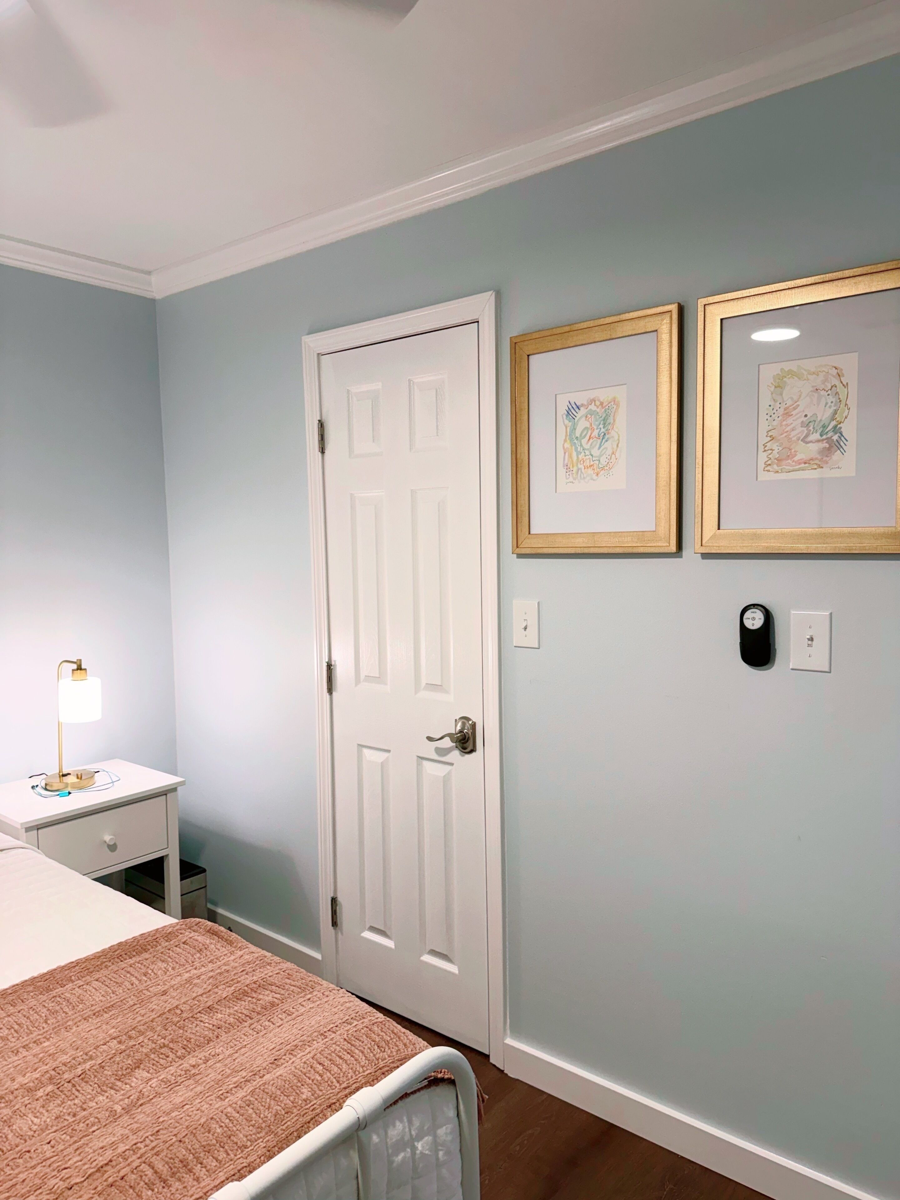 A bedroom with a bed, bedside table, lamp, door, and two framed pictures on the wall.