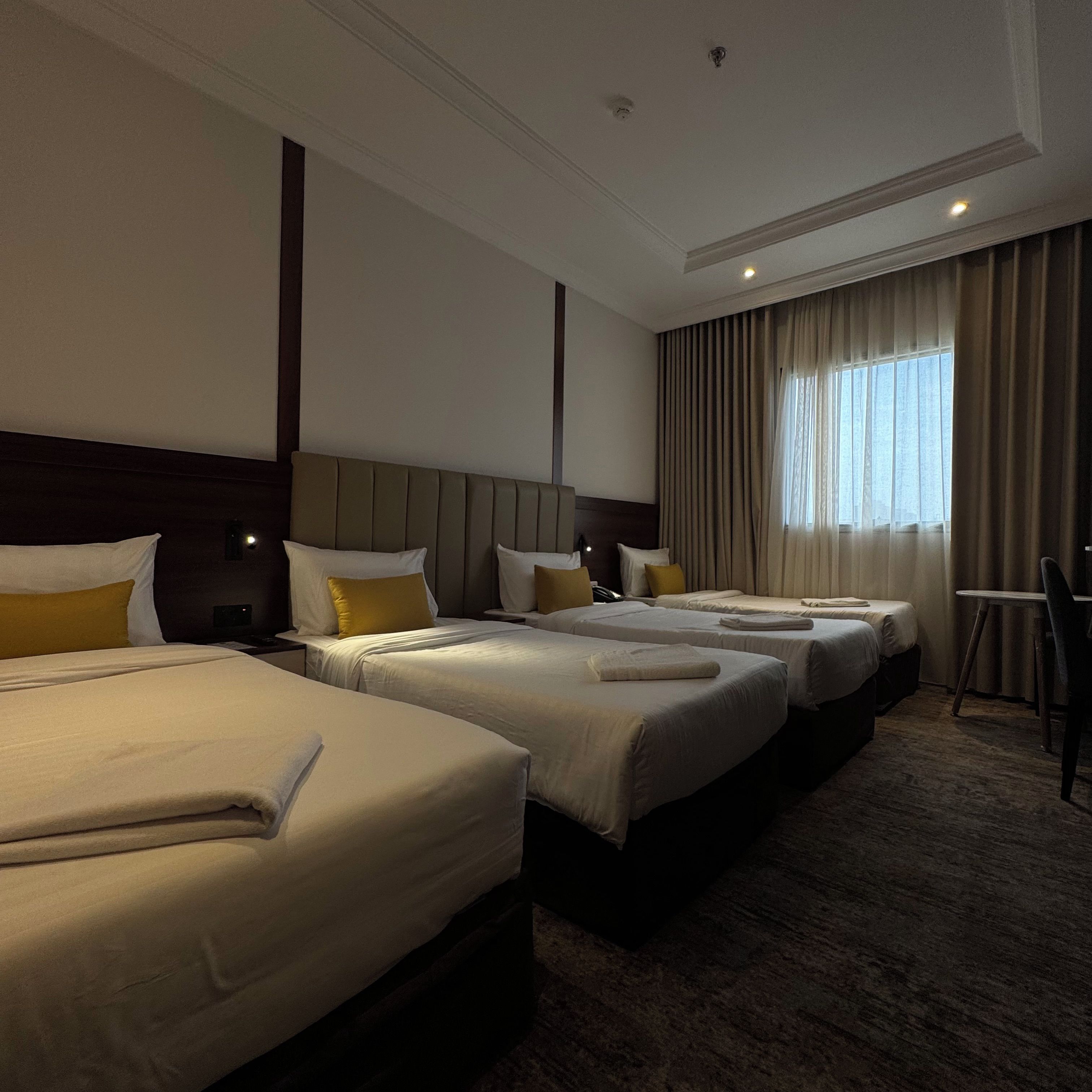 Habitación de hotel con cuatro camas dispuestas en fila, cabecero de madera, ventana grande con cortinas y una pequeña mesa con una silla.
