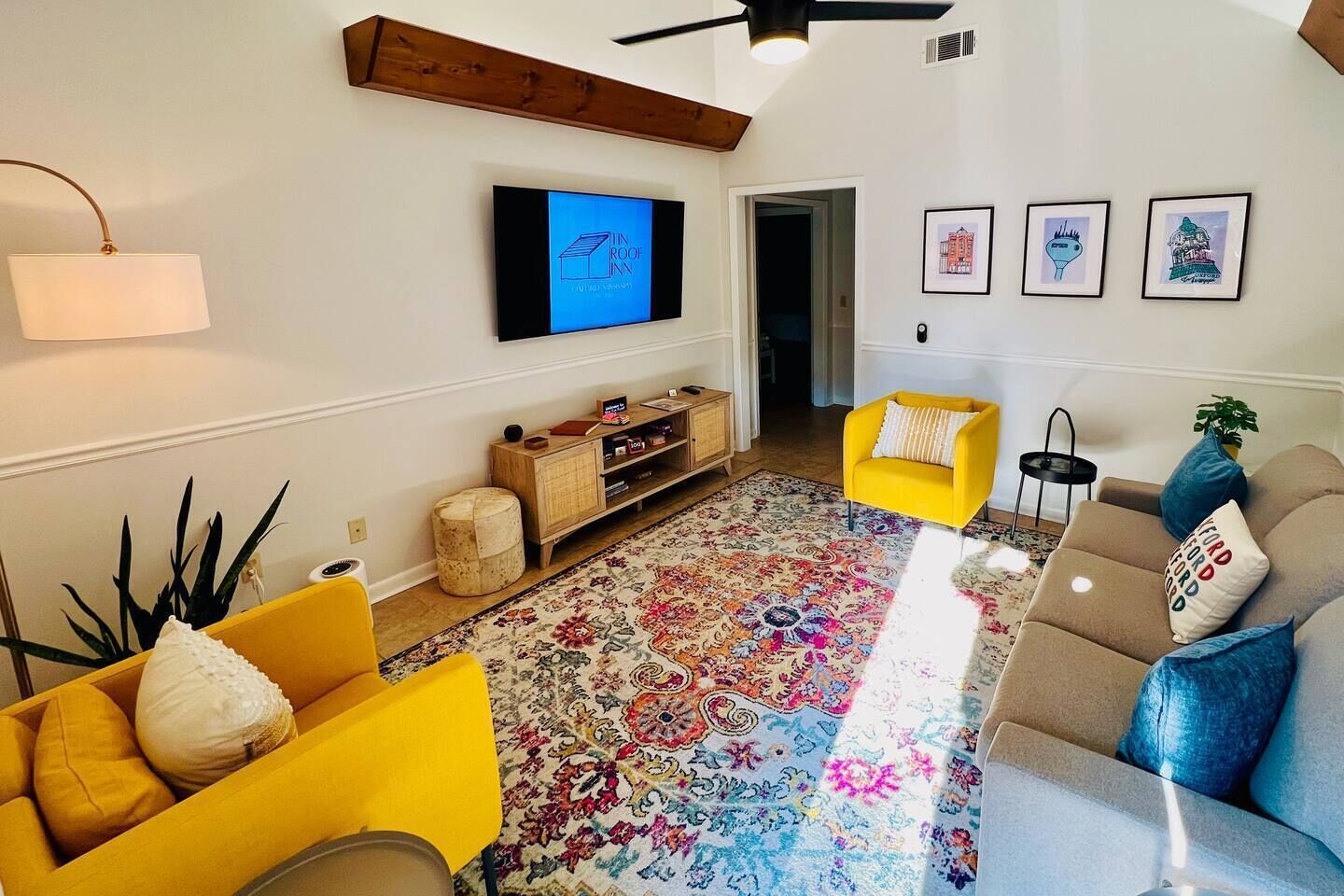 Ein Wohnzimmer mit Fernseher, gelben und grauen Sofas, einem gemusterten Teppich und gerahmten Bildern an der Wand.